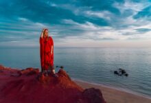 A Young Iranian Woman Stands On The Red Cliffs Of Hormuz Island, Where Vivid Earth Meets The Calm Waters Of The Persian Gulf.